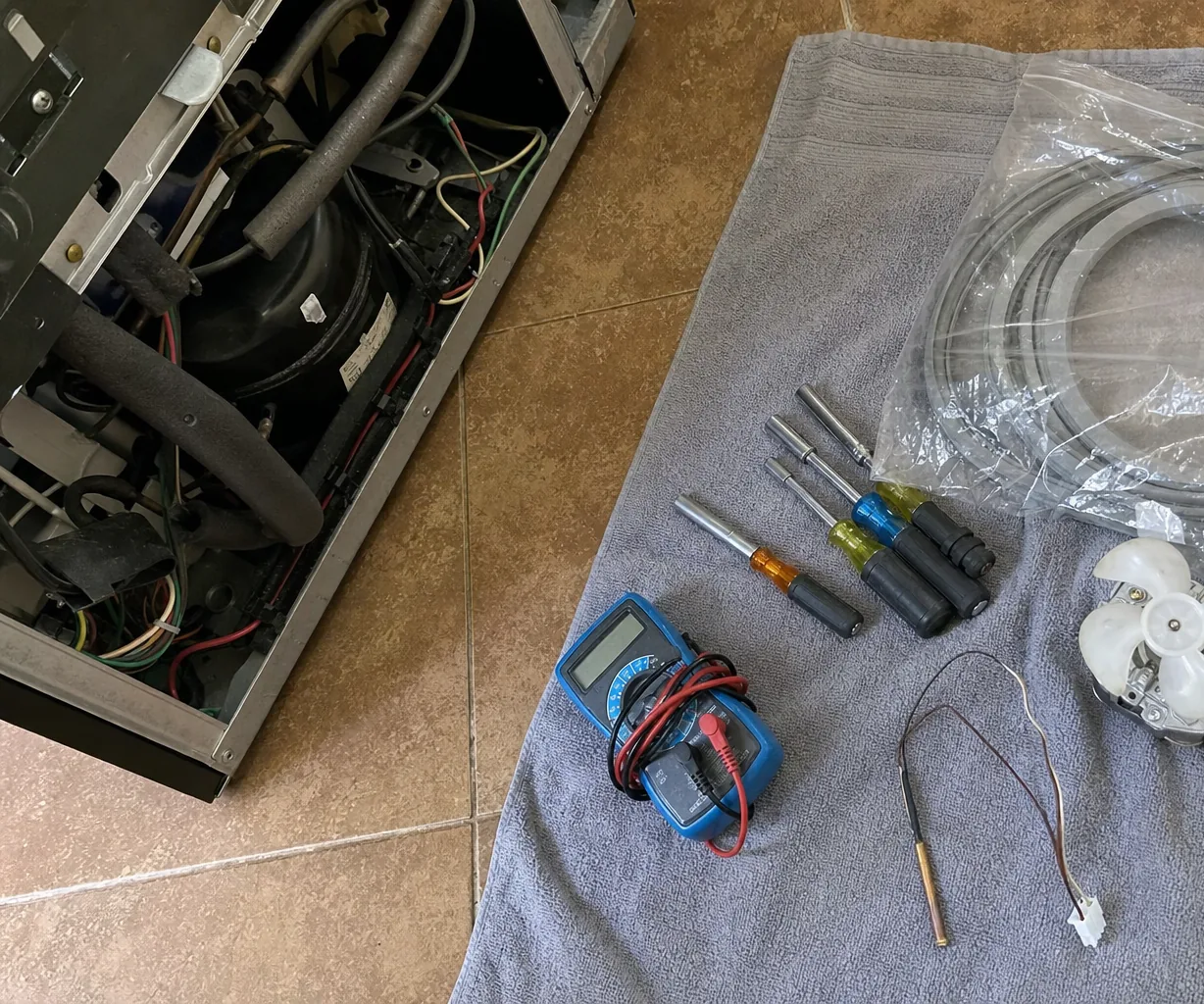 Left-hand view of a service kit spread on a towel on a tiled floor beside an appliance opened at the back